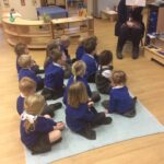 young pupils sitting on a mat in the classroom, with teacher showing them a page from a book